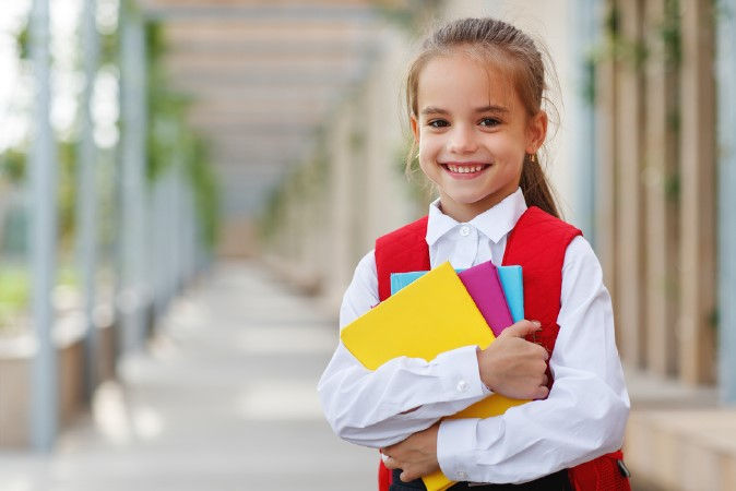 Smiling girl with a red backpack holds colorful books in a bright, open hallway. She's wearing a white shirt, conveying a cheerful mood.