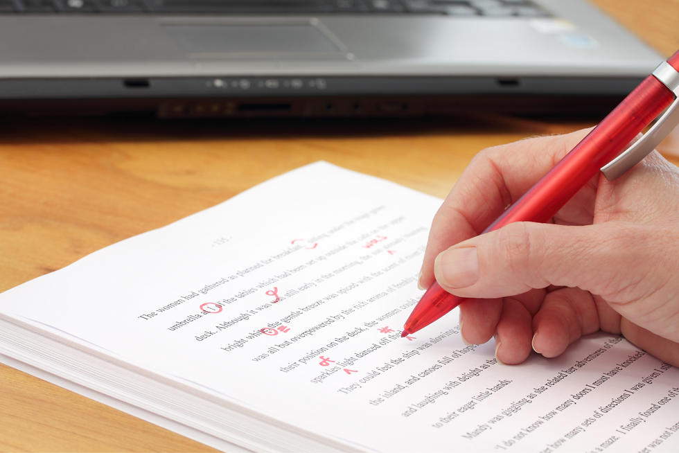 Hand editing a printed document with a red pen on a wooden desk; a laptop partially visible in the background.