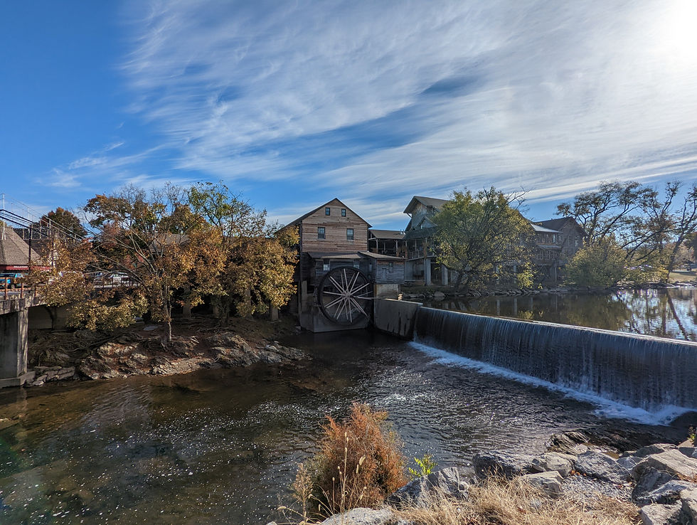 Old mill with waterwheel by a flowing river. Autumn trees surround the wooden buildings under a bright blue sky with wispy clouds.