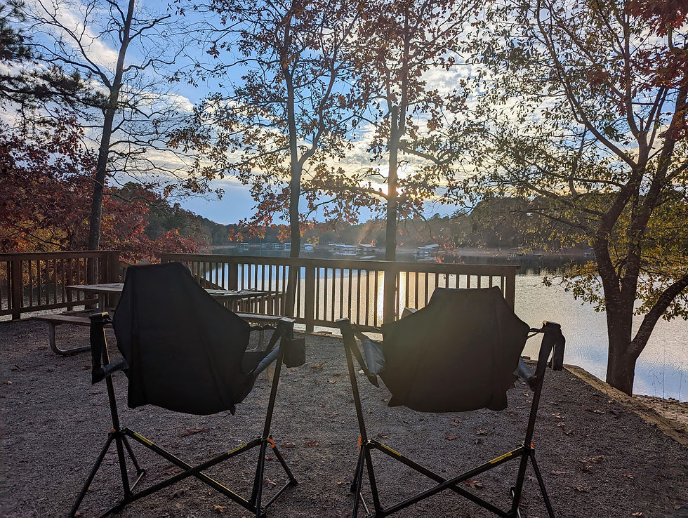 Two empty black chairs face a lake at sunset. Trees with autumn leaves surround a wooden deck. Peaceful, natural setting.