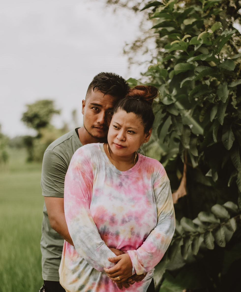 Latino man holding woman from behind and leaning on her. They stand in a field and the man looks at the camera.