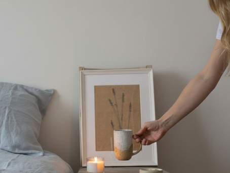 Femme souriante posant une tasse bicolore sur une table de chevet, près d’une bougie et d’un cadre dans une chambre.