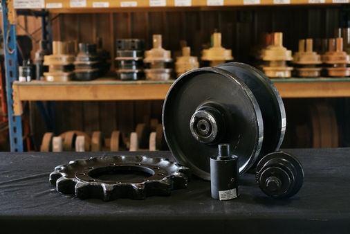 Sprocket, top roller or carrier roller, idler and bottom roller for mini excavator in front of a warehouse shelf with top rollers.