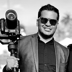 Alt text: Black and white photo of a man in a suit holding a professional camera with a stabilizer, possibly at an outdoor event such as a wedding. The man's face is blurred for privacy. He stands next to a woman dressed in dark attire, partially visible on the right. The background shows blurred outdoor elements, including chairs and palm trees, indicating a formal occasion or ceremony.