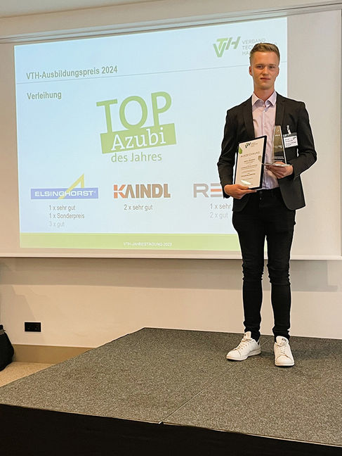 Ein Mann mit einer Auszeichnung in der Hand vor einem Bild mit der Aufschrift "Top Azubi des Jahres"