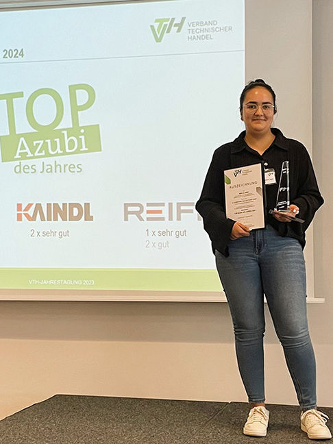 Eine Frau mit einer Auszeichnung in der Hand vor einem Bild mit der Aufschrift "Top Azubi des Jahres"