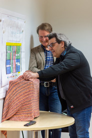 Zwei Männer die an einem Tisch stehen. Auf dem Tisch steht ein Gegenstand der mit einem Tuch abgedeckt ist.