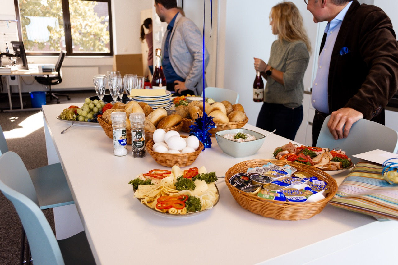 Ein Frühstücksbuffet auf einem Tisch. Hinter dem Tisch stehen drei  Personen.