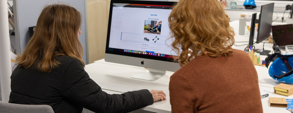Zwei Frauen die an einem Schreibtisch sitzen und auf einen PC-Bildschirm schauen.
