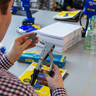 Einer Person die etwas auf ein Werkzeug stehen möchte. Im Hintergrund ein Tisch mit weiteren Werkzeugen und Collegeblöcken.