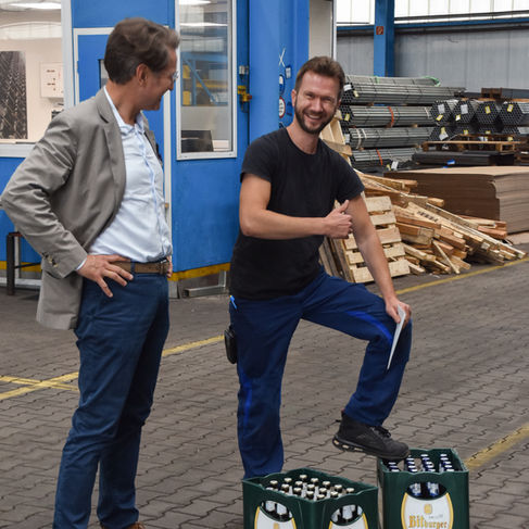 Zwei Männer stehen in einer Stahlhalle. Einer von den Männern hat seinen Fuß auf einem Kasten Bier stehen und zeigt einen Daumen nach oben.