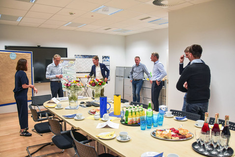 In einem Raum sieht man einen gedeckten Tisch. Im Hintergrund stehen mehrere Personen.