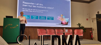 Short-haired woman in dark trousers and bright top, standing on stage next to a podium. Large screen with text and an ullustrative image of a laptop behind her. Row of chairs. 3D printed letters of SWEDMA standing on the floor in the foreground.