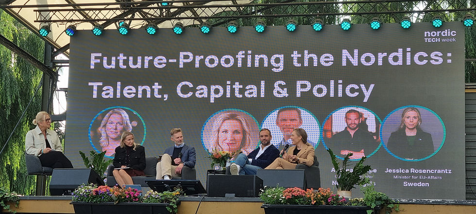Five people sitting on a panel, microphones in hand,  on a stage outdoors. A large backdrop screen has a title of the panel topic (Future-Proofing the Nordics: Talent, Capital & Policy) and pictures, name and title of the panelists.  Event logo in the upper right-hand corner. Flower beds in the foreground. Colored spotlights and a roof above the stage.
