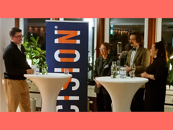 Four people (two men and two women) at standing tables with a Cision roll-up and some windows at the back.