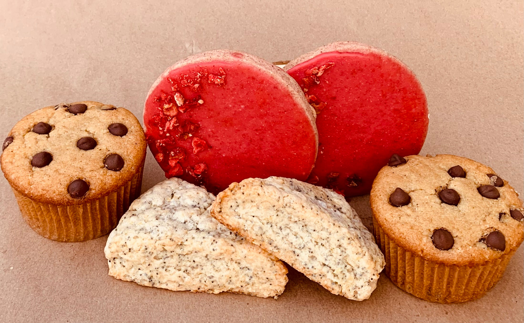 Two strawberry shortbread cookies standing on end behind two lemon poppy seed scones flanked by chocolate chip muffins