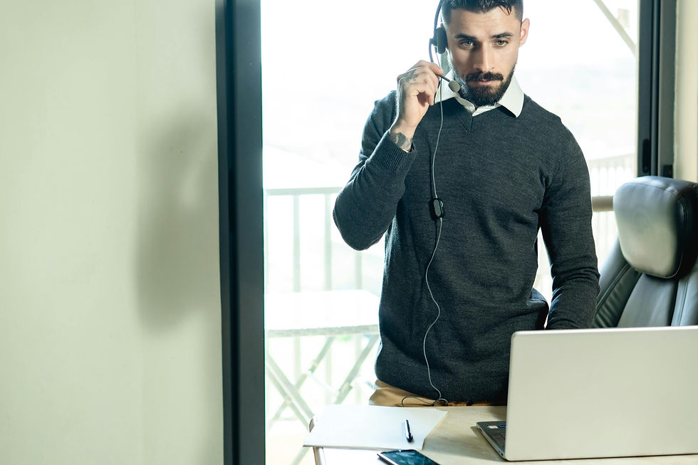 Man with headset at desk, holding mic, focused expression. Laptop, phone, and papers are on the table. Bright room with window view.