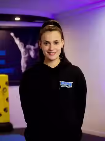 Makayla Giovaniello, instructor at Mozhdeh Martial Arts, smiling in a black MMA hoodie, standing in front of a gym wall with martial arts graphics