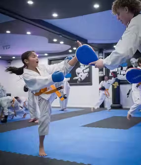 Child developing sparring skills by kicking pad held by instructor at Mozhdeh Martial Arts
