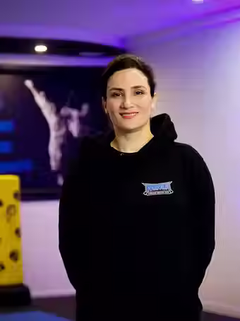 Shahnaz Mozhdeh, instructor at Mozhdeh Martial Arts, wearing a black MMA hoodie, standing in the dojo with a gentle smile