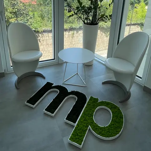 Logo MP realizzato con muschio stabilizzato in tonalità verde scuro e chiaro, esposto in un ambiente moderno e luminoso.