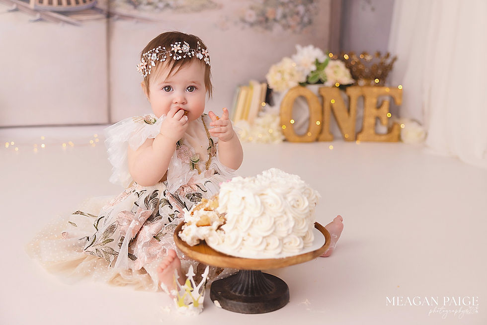 Baby in floral dress eating cake, sitting on the floor. Decorative "ONE" letters and flowers in background. Playful, celebratory scene.