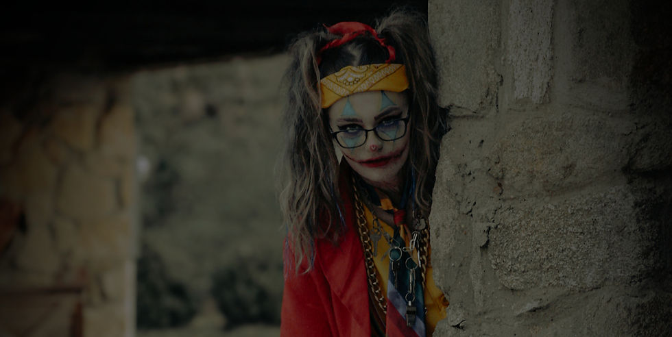 Woman in clown makeup, red jacket, and yellow bandana peeks around a stone wall. Dark, eerie setting with a mysterious mood.