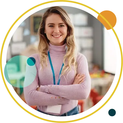 Smiling woman in pink turtleneck with lanyard stands arms crossed in a blurred classroom or office.