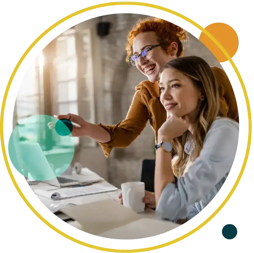 Two people smile while working at a desk with a laptop. One points at the screen, the other holds a mug. Bright, modern office setting.