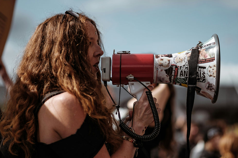 Woman with long wavy hair speaks into a sticker-covered megaphone at an outdoor protest under a blue sky.