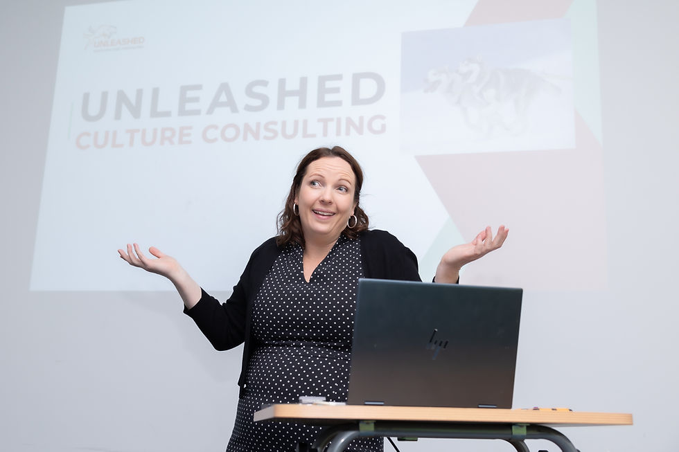 Woman at podium shrugs beside laptop during a talk, with projected slide reading UNLEASHED CULTURE CONSULTING in the background.