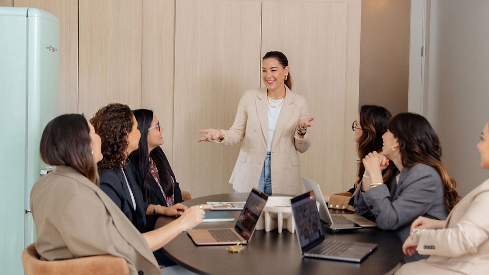 Smiling woman in a beige blazer presents to a group of women around a meeting table with laptops in a bright office.