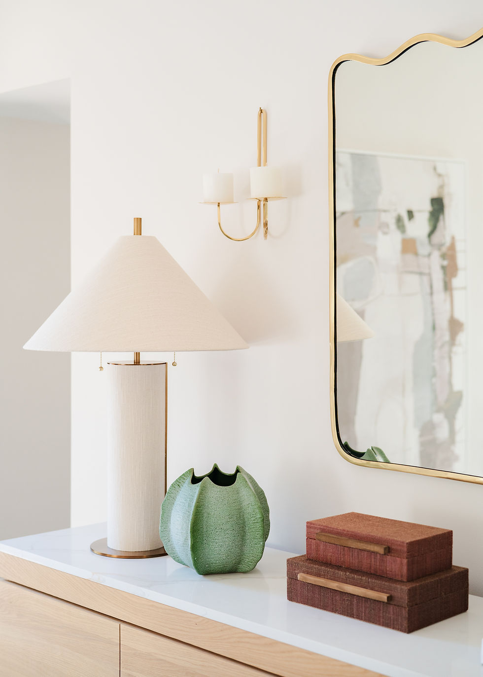 Elegant table setup with a beige lamp, green vase, and stacked boxes. Gold-framed mirror and candle fixture on white wall. Minimalist decor.