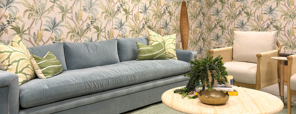 Cozy tropical lounge with rattan chairs, green pillows, framed beach photos, and leafy wallpaper in a bright, calm setting