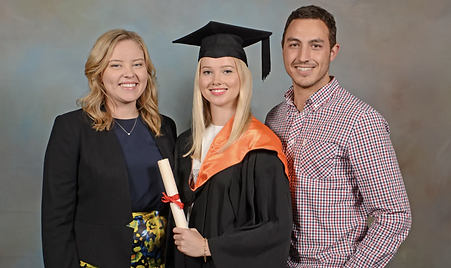 A graduation studio portrait of three smiling people. A blonde woman in a black cap, gown, and orange hood holds a diploma, flanked by a woman in a black blazer and a man in a checkered shirt.