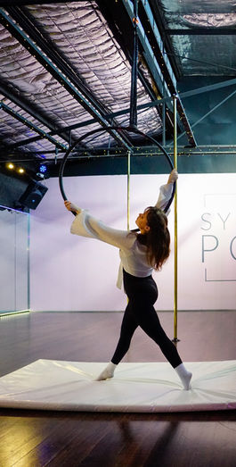 A photograph from the Sydney Pole series capturing a live showcase night, featuring an aerial dancer performing against a backdrop of industrial textures and vibrant pink lighting.