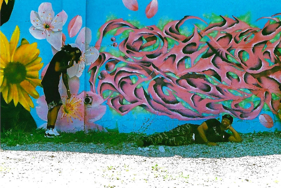 alt="Artists Propa posing in front of his pink flowers. While Apeks relaxes on the ground in front of his graffiti in Grove Hall"