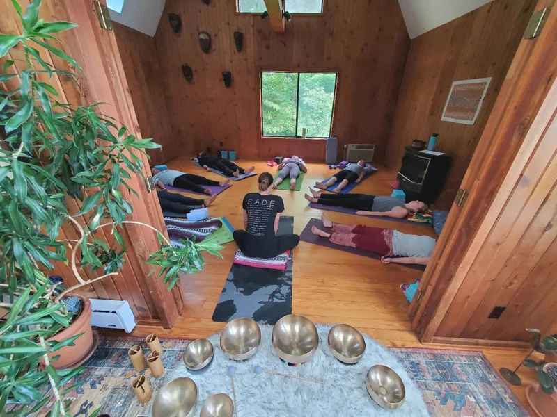 In a large wood framed room, eight people lay on yoga mats in a circle, with Anjali facing them. Behind Anjali is a set of singing metal bowls on a sheepskin rug
