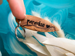 Schlüssel Anhänger in Surfbrett Form mit "paradise ♥️ " Gravur. Im Hintergrund Meeresblau und getrocknete helle Blumen.