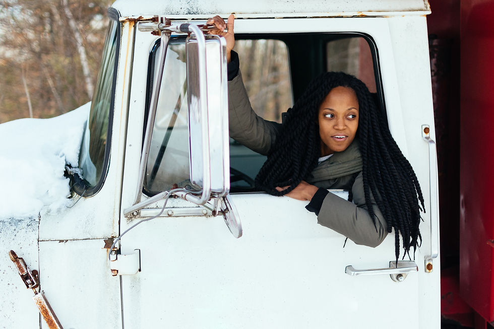 A black woman leans out of a large truck's window, looking back and smiling