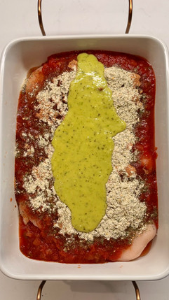 Red and green salsa and seasonings covering raw chicken breasts in a white baking dish with brass handles on a white countertop
