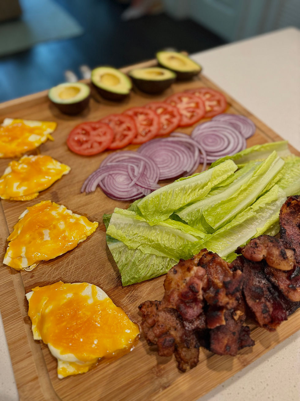 Avocado halves, tomato and onion slices, lettuce, eggs with melted cheese, and bacon on a wooden board, ready for sandwich assembly.