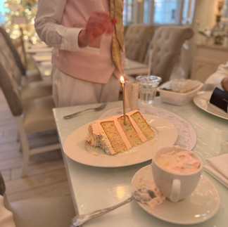 A large slice of white cake with sprinkles and fluffy icing with a gold candle lit in the middle of it. A waitress in the background of a fancy French restaurant