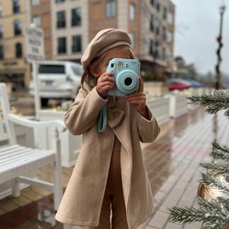 A little girl wearing brown winter jacket and French hat holding a blue Polaroid camera up to her face taking pictures