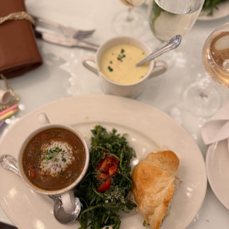 On a white table sits a cup of creamy chicken soup and a cup of gumbo soup topped with white rice and fresh herbs. A mixed green salad with a chicken salad crossiant next to it on the palte