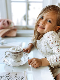 Little girl wear a white lace dress with a tea cup full of hot chocolate topped with marshmallows and whipped cream topping.