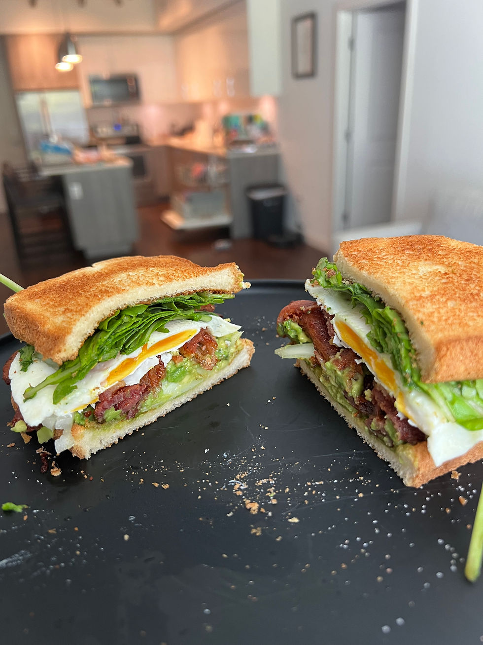 Two BLT sandwiches with lettuce, bacon, avocado, and egg on toasted bread sit on a black plate in a kitchen with light cabinets.