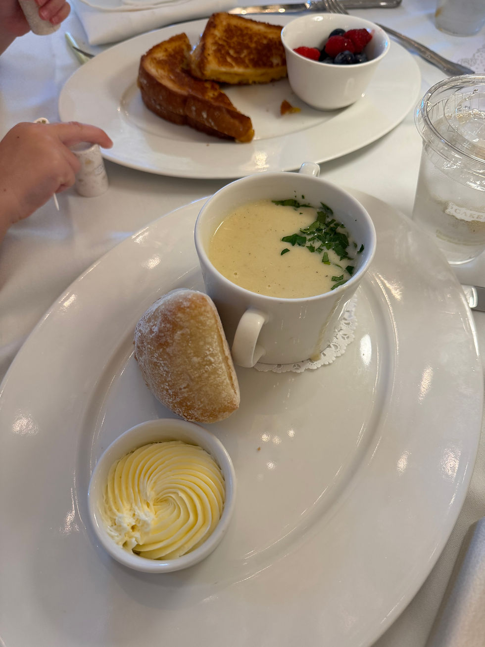A plate with soup, bread, and butter, alongside French toast, berries, and a child's hand near salt. White table setting, bright mood.