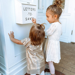 Two little girls putting paper into a white box that reads letters to Santa. Girls are wearing a white lace dress and a gold metallic dress with stockings