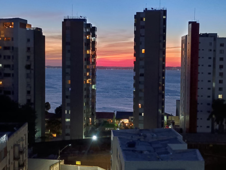 A view of the sea with a pink-and-orange tinged sky is partially obstructed by apartment buildings sticking up like dominoes or dolmens, their lights slowly coming on in the twilight.